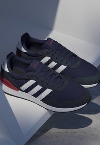 Par de zapatillas Adidas azul marino con rayas blancas y detalles rojos sobre una superficie blanca inclinada.
