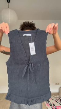 Persona con cabello rizado sostiene una blusa sin mangas de color gris oscuro con textura, nudo frontal y dobladillo con volantes en interiores.