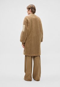 Brūns, pūkains vilnas kažoks ar pārlieku lielu piegriezumu, lielām piedurknēm un bēša auduma akcentiem plecos. Komplektā ar vaļīgām bēša krāsas biksēm.