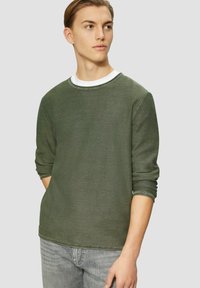 Sweatshirt vert à col rond avec des manches longues, fabriqué à partir d'un tissu texturé. Superposé avec une chemise blanche en dessous. Porté avec un jean gris clair.
