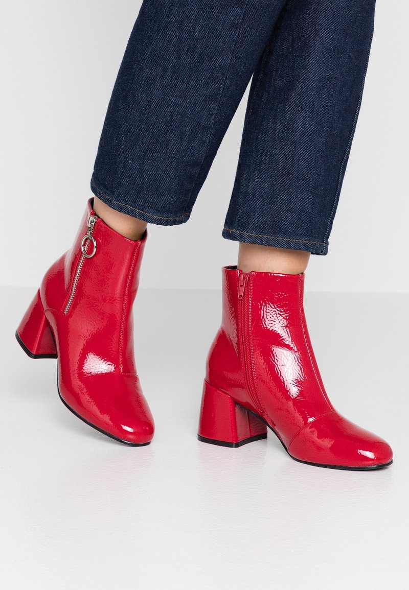 Bottines rouges brillantes en cuir verni avec talons carrés et fermetures éclair latérales portées avec un jean court bleu foncé sur un sol blanc.