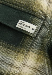Étiquette sur tissu à carreaux vert et beige avec l'inscription « PLAY without LIMITS Since Eighty Two » avec un petit symbole d'empreinte de patte.