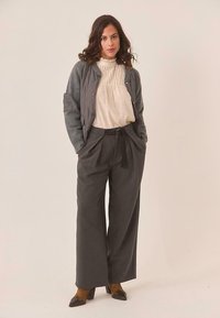 Chaqueta bomber acolchada gris sobre una blusa beige con pliegues, combinada con pantalones de pierna ancha gris oscuro y tacones marrones. Texturas suaves y ajuste a medida.