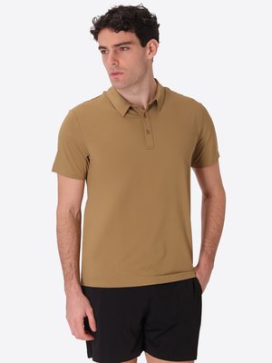 Uomo con capelli ricci scuri che indossa una polo marrone a maniche corte e pantaloncini neri, guarda verso sinistra con una mano in tasca.
