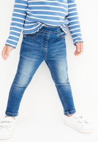 Modrá a biela prúžkovaná blúzka s dlhými rukávmi a riasenými manžetami, skombinovaná s priliehavými modrými džínsami a bielymi teniskami. Džínsové nohavice majú vyblednutý dizajn.