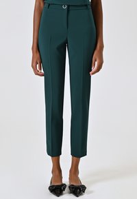 Grüne, maßgeschneiderte Hose mit glatter Textur, ausgestattet mit einem Gürtelschlaufen und einem metallischen Akzent. Der Saum sitzt über dem Knöchel.