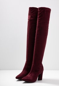 Botas de terciopelo burdeos hasta la rodilla con punta afilada y tacón grueso, con una textura lisa y elegante y sin adornos visibles.