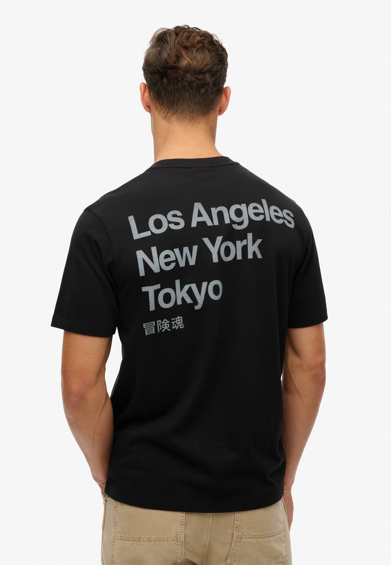 Camiseta de algodón negra con texto en gris llamativo en la parte posterior: "Los Ángeles," "Nueva York," "Tokio," con caracteres chinos debajo.