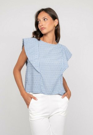 Femme aux cheveux foncés portant des boucles d'oreilles en or, un haut sans manches à carreaux bleu et blanc avec de grands volants, et un pantalon blanc les mains dans les poches.