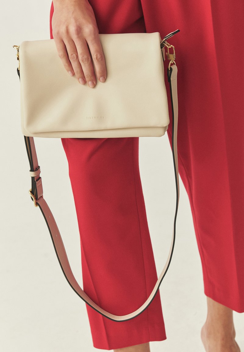Hand hält eine beige Leder-Clutch mit langem Riemen, getragen über roten, maßgeschneiderten Hosen.