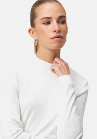 White long-sleeve top with a high neck, featuring a smooth texture. Model touches collar while wearing large silver hoop earrings.