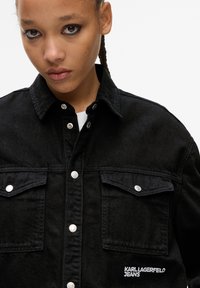 Black denim shirt with two front pockets, silver snap buttons, and embroidered "KARL LAGERFELD JEANS" logo on the left side.