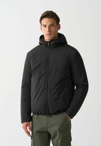 Schwarze isolierte Jacke mit Kapuze, ausgestattet mit einem Reißverschluss auf der Vorderseite und strukturiertem Stoff. Das Model trägt olivgrüne Cargo-Hosen und steht vor einem weißen Hintergrund.