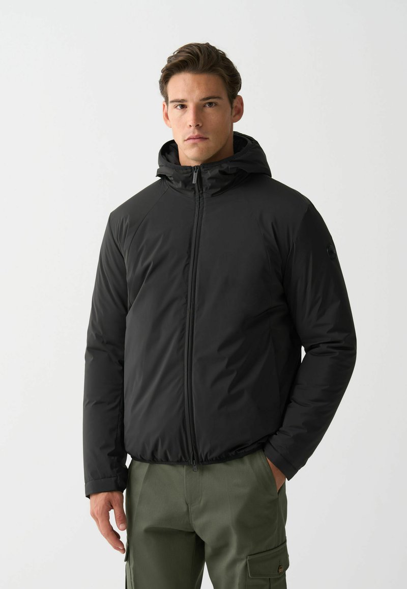 Schwarze isolierte Jacke mit Kapuze, ausgestattet mit einem Reißverschluss auf der Vorderseite und strukturiertem Stoff. Das Model trägt olivgrüne Cargo-Hosen und steht vor einem weißen Hintergrund.