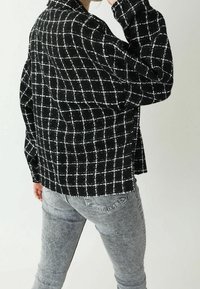 Person som bär en svartvit rutig oversize-jacka och blekta grå jeans, poserar lite vänd bort med handen nära ansiktet.