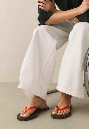 Braune Flip-Flops mit leuchtend roten Riemen. Person sitzt im Rollstuhl, trägt weiße Hosen mit weitem Bein und ein schwarzes Oberteil. Glatte Textur und minimalistisches Design.