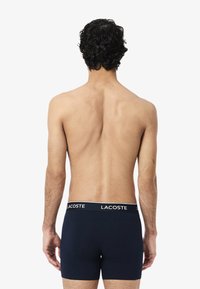 Námornícky modré boxerky s bielym pásom, na ktorom je logo Lacoste. Strih je priliehavý, zdôrazňuje hladkú textúru a čisté línie.