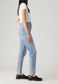 Kvinna i ljusblå hög midja jeans, vit kortärmad topp och svarta loafers som står mot en enfärgad bakgrund.