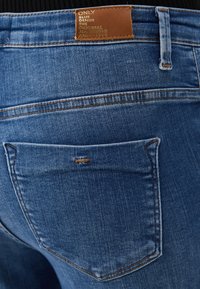 Jeans de mezclilla azul con una etiqueta de cuero en la cinturilla. Incluye un bolsillo trasero cosido con costuras visibles y detalles de textura.