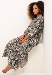 Abito lungo e largo in beige con strisce zebra nere, caratterizzato da un collo rotondo, maniche lunghe e spacchi laterali. Tessuto dalla texture liscia.
