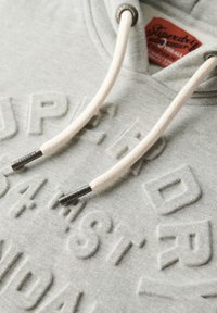 Sudadera gris con letras en relieve texturizadas de "Superdry". Presenta cordones blancos con acentos metálicos y una etiqueta en el cuello.