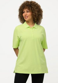 Donna con capelli ricci, indossa una polo a maniche corte di colore verde lime e pantaloni neri, in piedi con le mani dietro la schiena, guarda verso destra.