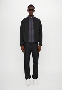 Herrenoutfit mit einer dunklen Jacke mit strukturiertem Finish, einer grauen Steppweste, dunklen Jeans und weißen Turnschuhen. Sonnenbrille inklusive.