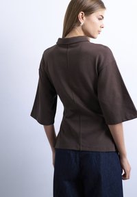 Ženska, oblečena v temno rjavo kratko-sleeve majico s ovratnikom in temno modre hlače, prikazana od zadaj na preprostem svetlem ozadju.