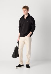 Čierna mikina na zips s vysokým golierom, béžové nohavice a čierne tenisky. Modelka nesie čiernu tašku cez rameno. Minimalistický dizajn.