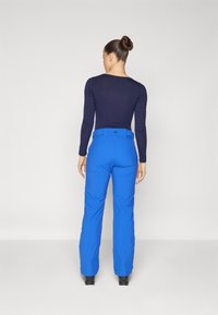 Μπλε παντελόνια σκι με λεία υφή και ελαφρώς φαρδιά γραμμή. Διαθέτουν ζώνη μέσης με λογότυπο, πλαϊνές τσέπες και ενισχυμένα μπατζάκια. Μπλούζα με μακριά μανίκια σε ναυτικό μπλε.