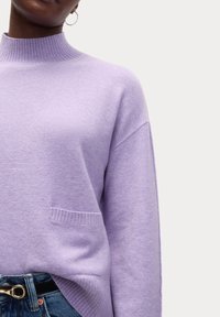 Lavendelfarbener Strickpullover mit hohem Rippkragen, überschnittenen Schultern und einer aufgesetzten Tasche. Der Stoff wirkt weich und hat eine feine Struktur.