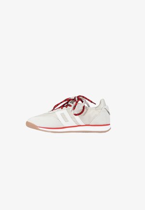 White low-top sneaker with red and black laces, beige suede heel, red stripe on sole, and white geometric side detailing.