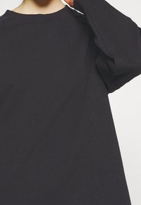 Svart långärmad skjorta med rundad krage, tillverkad av bomull, med avslappnad passform och mjuk textur. Detaljerad sömnad synlig på axeln.