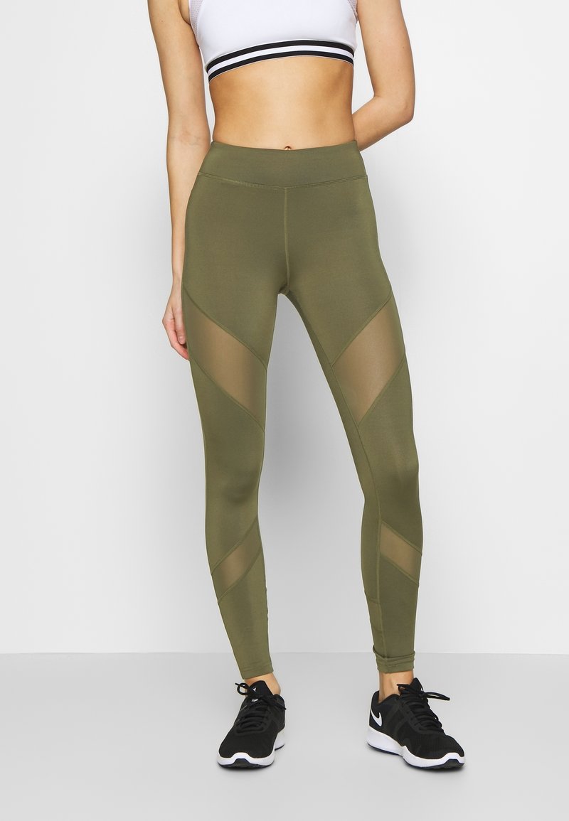 Olivgröna leggings med nätpaneler i ett diagonalt mönster, tillverkade av stretchigt material. Matchas med svarta träningsskor som ger en åtsittande look.