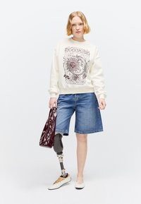 Jeune femme aux cheveux blonds courts portant un sweat-shirt graphique, un short en jean, des ballerines blanches et une jambe prothétique, tenant un sac bordeaux clouté.