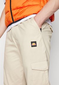 Shorts cargo beiges avec un patch noir arborant un logo coloré. Les shorts ont des poches latérales et une coupe droite.