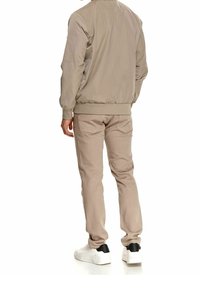 Homme portant une veste bomber beige, un pantalon beige assorti et des baskets blanches avec un détail noir à l'arrière, se tenant de dos à l'appareil photo.