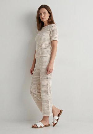 Vrouw staande in een crèmekleurige gehaakte top met korte mouwen en bijpassende broek met witte platte sandalen tegen een simpele lichte muur.