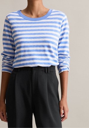 Persoon die een blauw-wit horizontaal gestreept shirt met lange mouwen draagt, ingestopt in een zwarte broek met hoge taille, met een effen achtergrond.