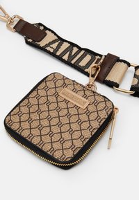 Borsetta zip con motivo geometrico nero su sfondo beige, accessori in oro e una tracolla staccabile con testo a contrasto. Materiale in tela morbida.