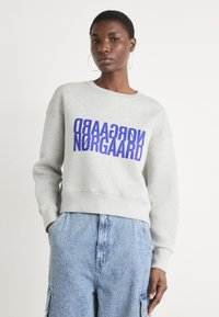 Grå hettegenser med rund hals, med fet lilla tekst "Nørgaard" trykt på forsiden; kombinert med teksturerte blå dongeri cargo-bukser.