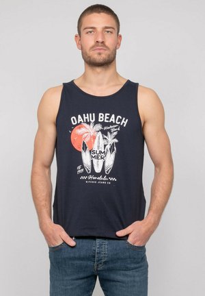 Jeune homme barbu portant un débardeur bleu marine avec inscription « Oahu Beach » et motif planche de surf, debout les mains dans les poches sur un fond uni.