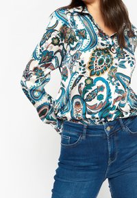 Kvinna klädd i en långärmad, knäppt blus med blå, gröna och orange paisleymönster, instoppad i blå denimjeans.