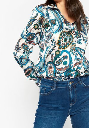 Kvinna klädd i en långärmad, knäppt blus med blå, gröna och orange paisleymönster, instoppad i blå denimjeans.