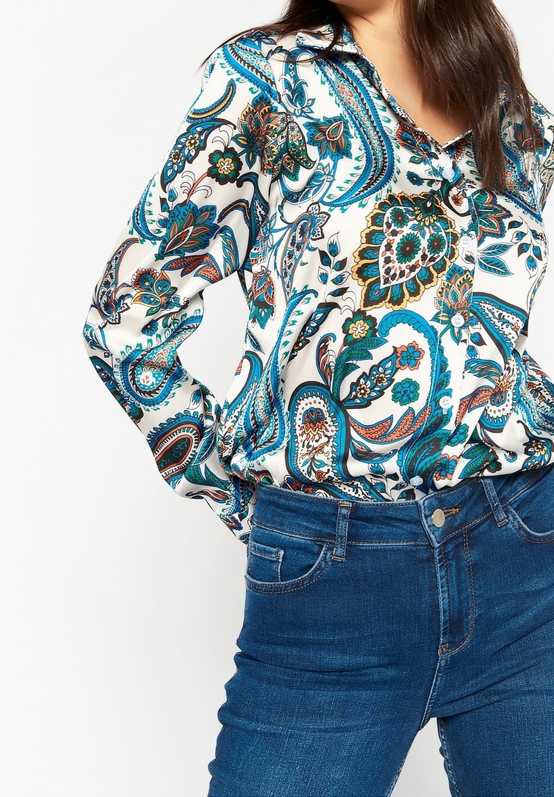 Kvinna klädd i en långärmad, knäppt blus med blå, gröna och orange paisleymönster, instoppad i blå denimjeans.