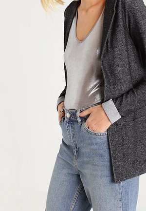 Blazer gris texturizado sobre una blusa metálica plateada, combinado con jeans de mezclilla azul de tiro alto. Destacan el escote en V y las mangas del blazer con puños.