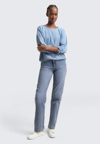 Lyseblå, teksturert skjorte med lange ermer og samlet mansjett, kombinert med rette blå jeans og hvite sneakers. En enkel, uformell antrekk.