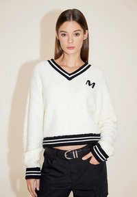 Weißer, flauschiger Pullover mit V-Ausschnitt und schwarzen Streifenakzenten am Kragen und Saum. Mit einem gestickten 'M'-Logo auf der Brust.