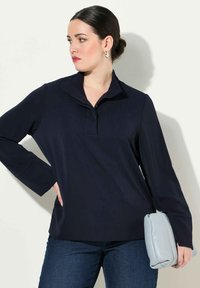 Navy lange mouw top met een opstaande kraag, gemaakt van zachte stof. Gecombineerd met blauwe jeans en een zachte grijze handtas.