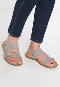 Sandales à glissière avec des brides argentées métalliques à finition texturée, posées sur une semelle beige ; design à bout ouvert mettant en valeur des ongles de pieds rouges polis.